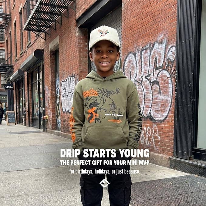 A young boy wearing a tan Mallard Duck Vintage Hat by Funky Junque and an olive green hoodie stands on a city sidewalk.