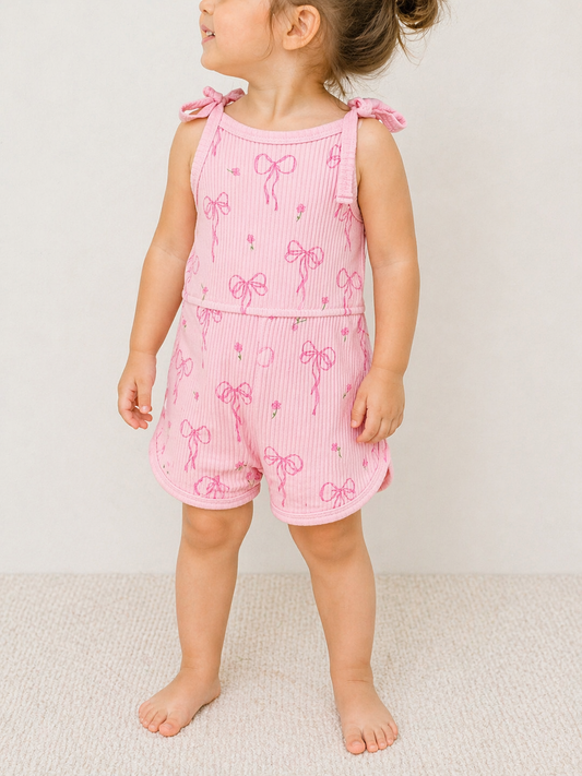 A young child stands indoors on a carpeted floor, wearing the Tiny Tot Bundles Pink Ribbon Tie Romper with bow patterns and tie straps, hair tied up, looking to the side against a neutral background.