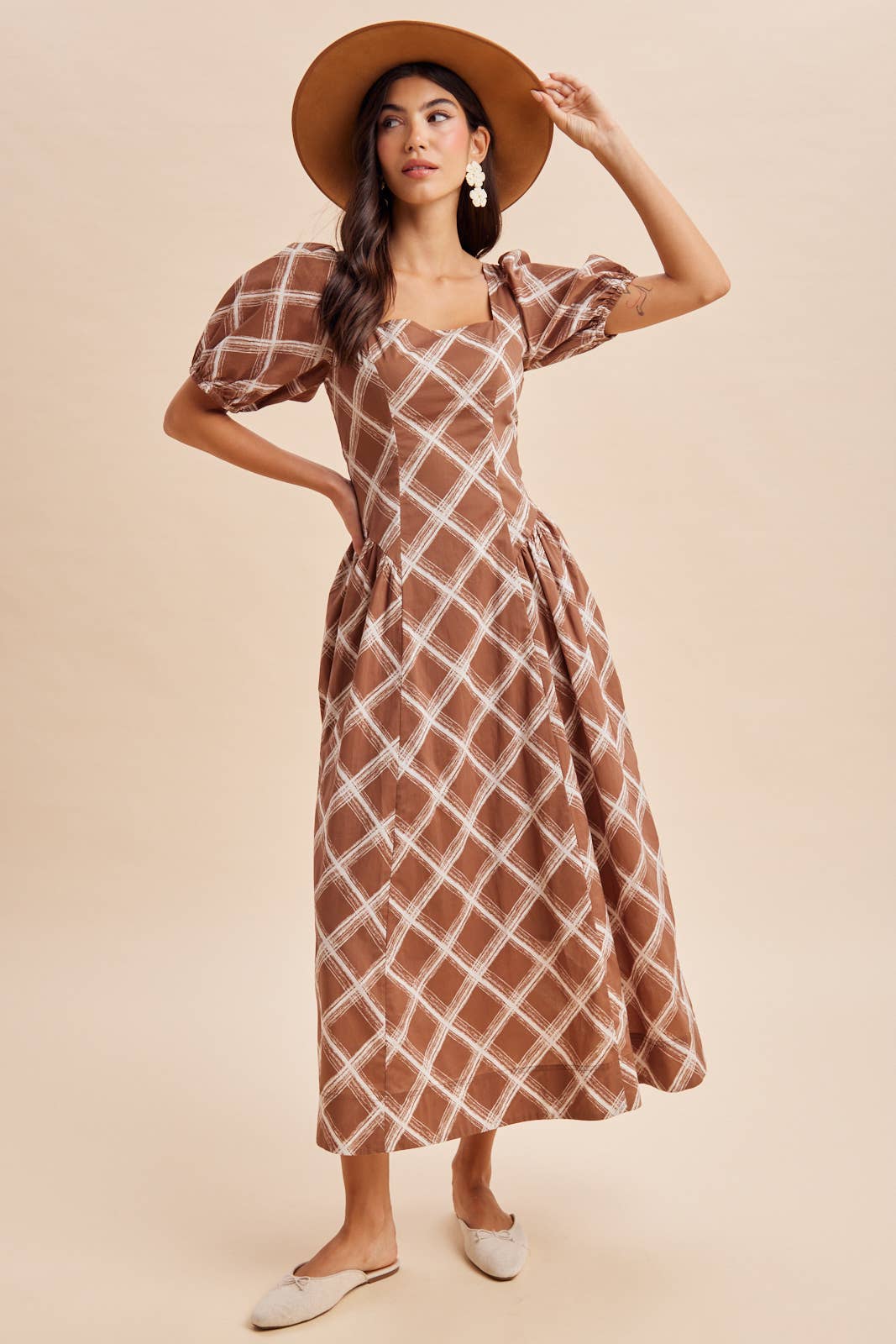 A person wearing an In Loom Plaid Square Neck Maxi Dress in brown and white with puff sleeves and a square neckline, paired with a wide-brimmed hat and flat shoes.