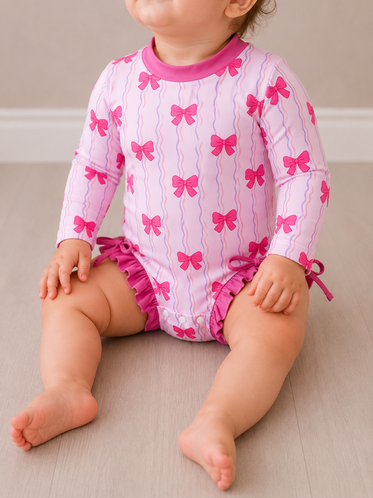 A baby sits on the floor wearing the Tiny Tot Bundles Pink Bow Swimsuit, a long-sleeve one-piece with wavy lines, pink bows, leg ruffles, and pink collar trim. The baby's face is not fully visible.