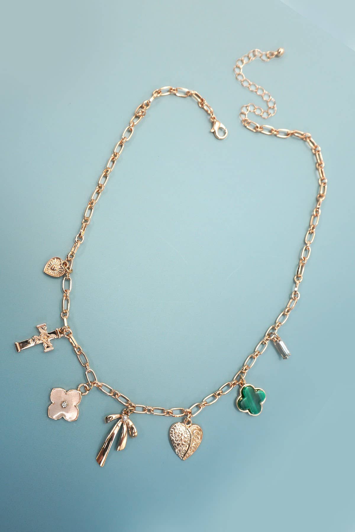 The Gold Charm Necklace by JOSSLYN by wall to wall features multiple gold pendants—a small heart, a cross, a flower, a person silhouette, a textured heart, a green clover, and a rectangular charm—elegantly displayed on a light blue surface.