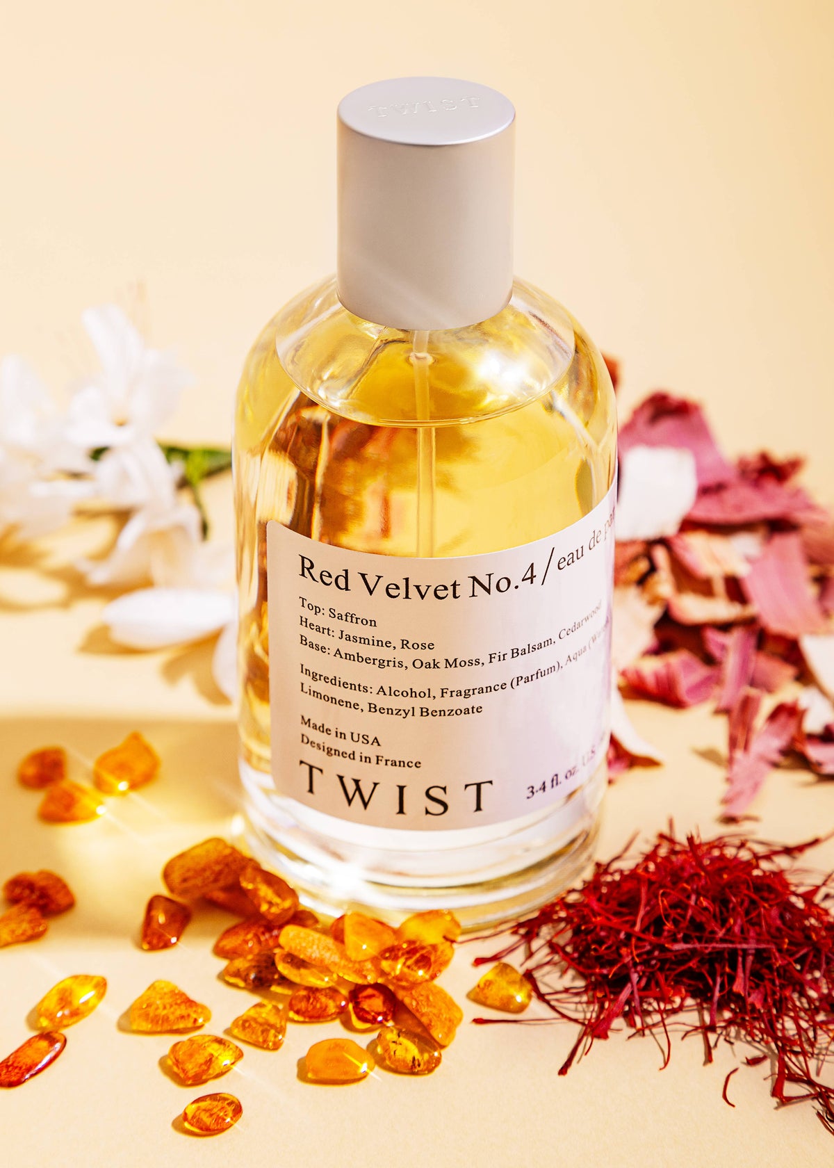 A bottle of House of Twist’s Red Velvet No. 4 Perfume—an elegant, long-lasting fragrance—is displayed among amber stones, saffron threads, and white flowers on a light beige surface. The label details its fragrance notes and ingredients, emphasizing the distinctive unisex character of this scent.