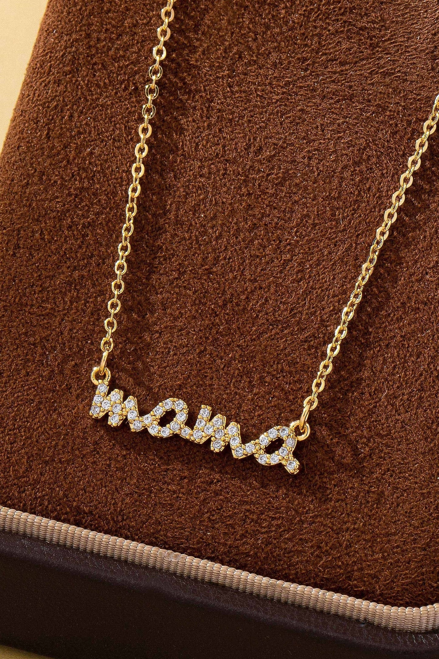 The JOSSLYN by wall to wall Gold MAMA Necklace features the word “mama” spelled out in cursive and encrusted with small sparkling stones, elegantly displayed on a brown velvet surface.