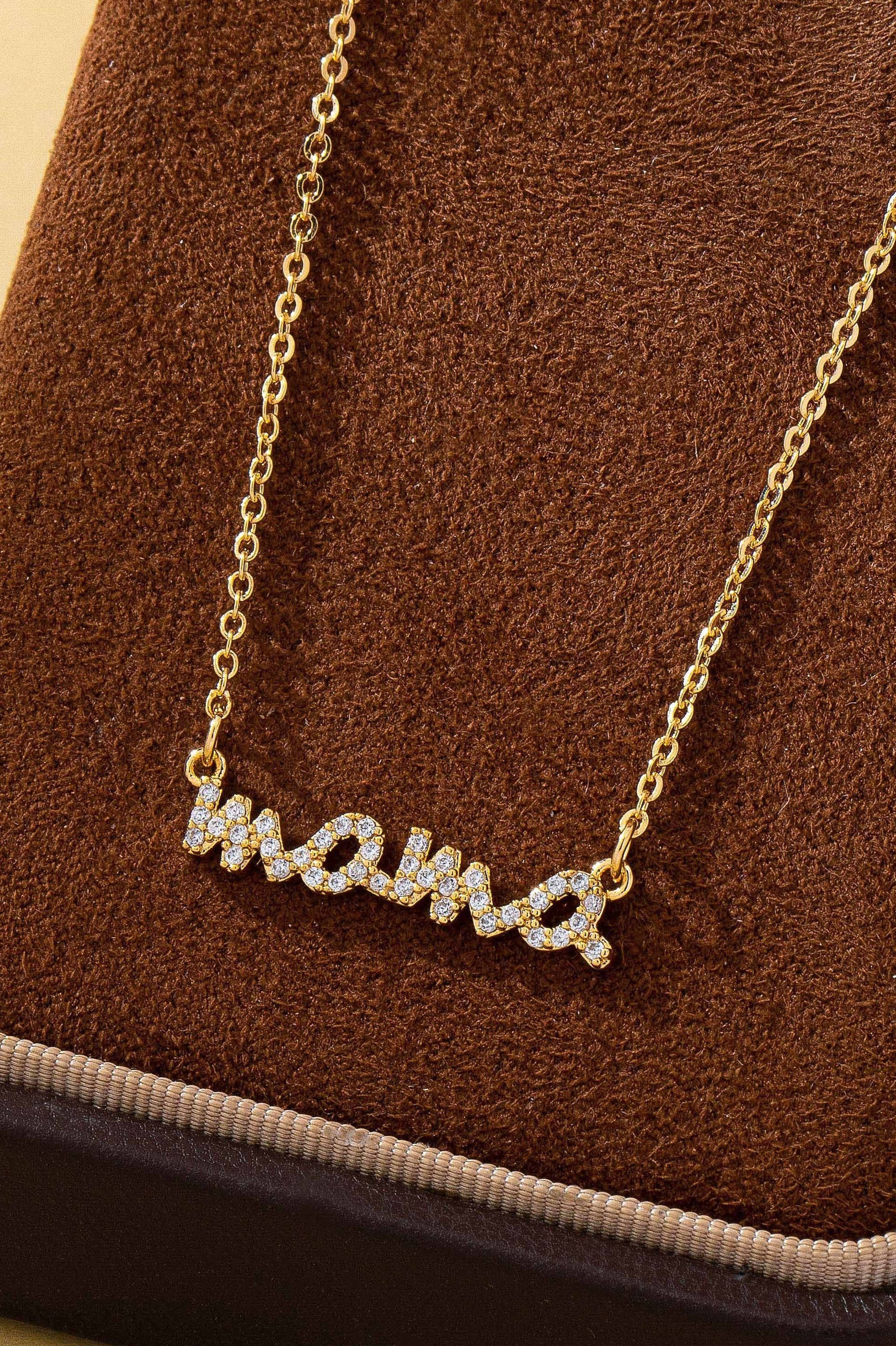 The JOSSLYN by wall to wall Gold MAMA Necklace features the word “mama” spelled out in cursive and encrusted with small sparkling stones, elegantly displayed on a brown velvet surface.