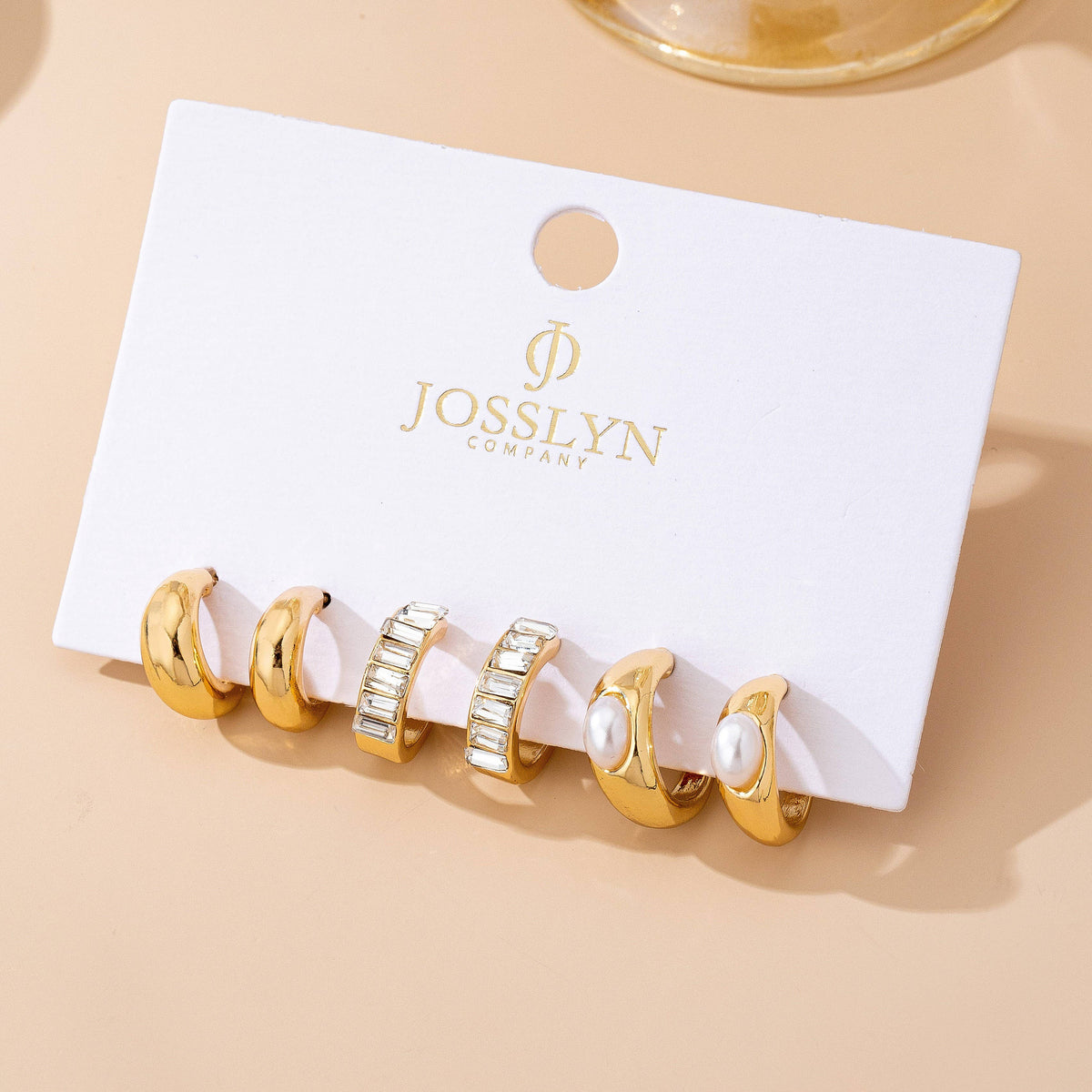 Five gold earrings from the Pearl & Baguette Hoop Trio Earring Set by JOSSLYN by wall to wall rest on a beige surface, displayed on a white branded card: two smooth hoops, two adorned with small crystals, and one featuring a pearl accent.