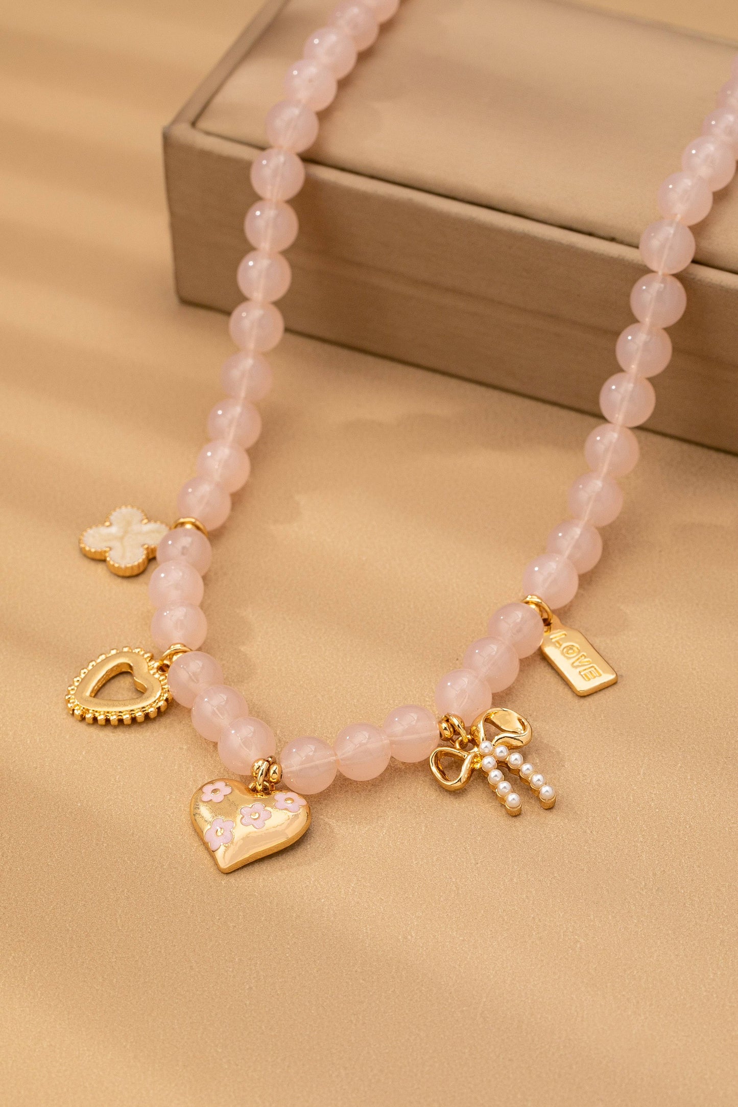 The Beaded Charm Necklace from JOSSLYN by wall to wall features pink beads with gold and pearl charms—hearts, a bow, a flower, and a rectangular tag—beautifully displayed on a beige surface alongside a tan box.