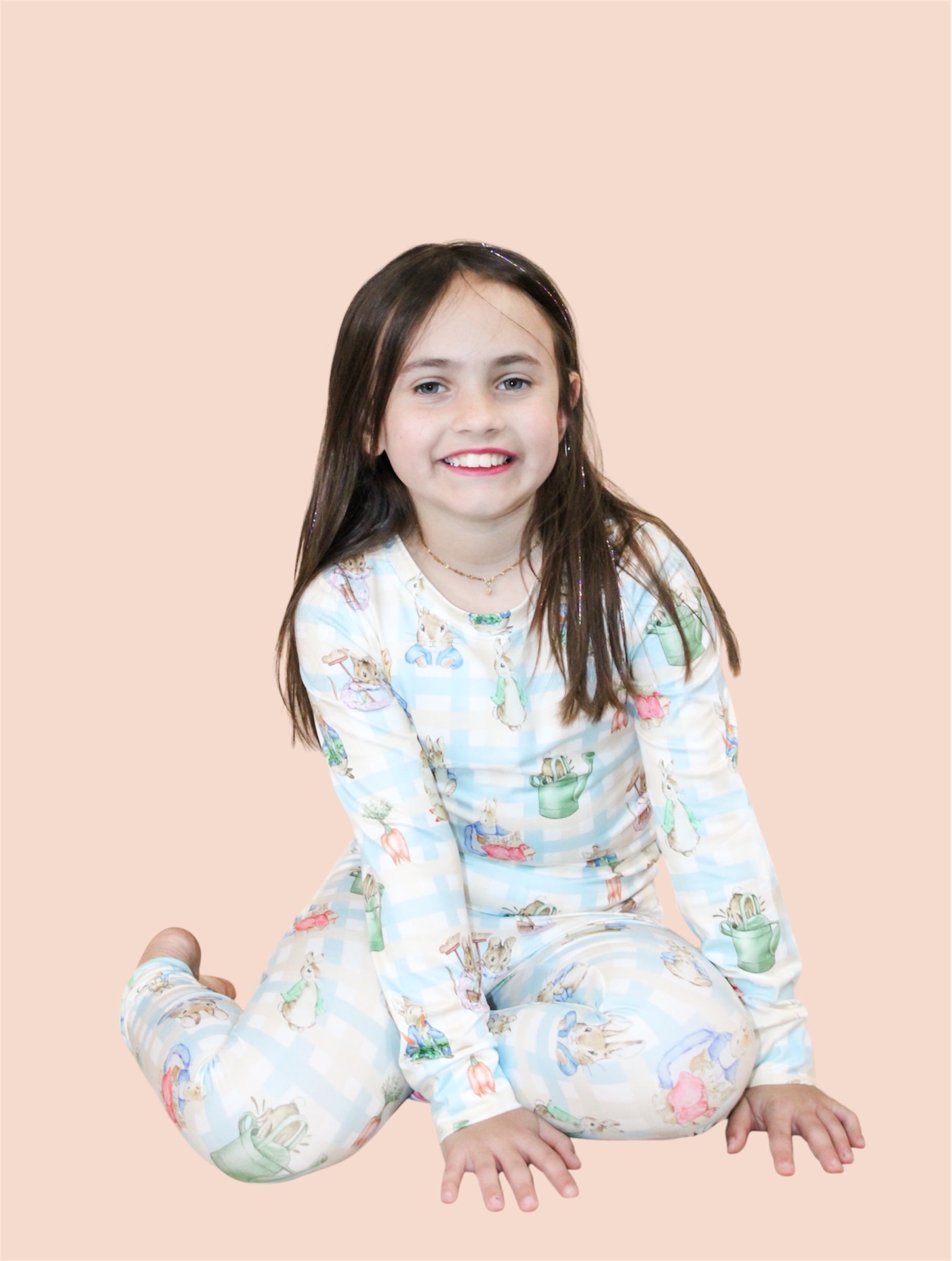 A young girl with long brown hair smiles while sitting on the floor in Tiny Tot Bundles' Peter's Easter Picnic Jammies Set, featuring playful animal prints, against a plain light peach background.