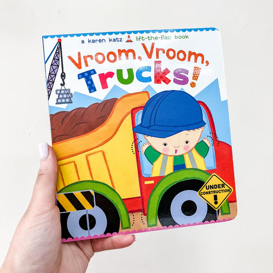 A hand holds the Vroom, Vroom, Trucks Book by Simon & Schuster, featuring a cover illustration of a smiling child in a hard hat driving a dump truck filled with dirt.