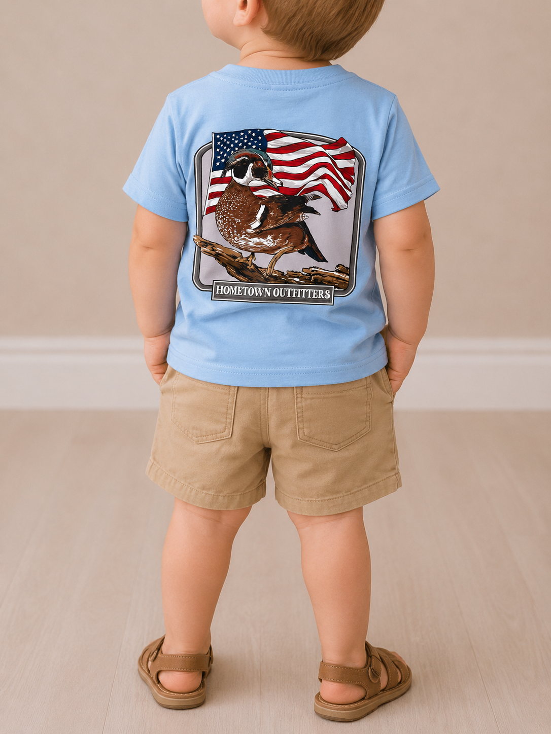 A young child stands indoors, wearing the Wild Threads Co Little Patriot T-Shirt featuring a quail and American flag graphic, along with tan shorts and brown sandals on a light wood floor.