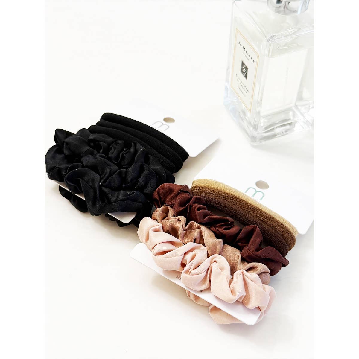 Two Love and Repeat Assorted Hair Tie & Scrunchie Sets, featuring black, brown, tan, and pink tones, are displayed on white cards next to a clear glass perfume bottle on a white surface.