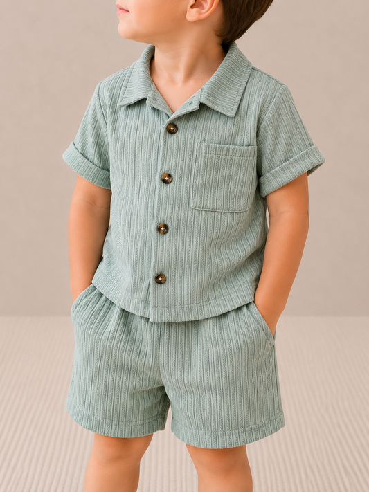 A young child models the Sage Breeze Set by Tiny Tot Bundles—light green, ribbed short-sleeve button-up shirt with pocket and matching shorts—standing with hands in pockets against a neutral background.