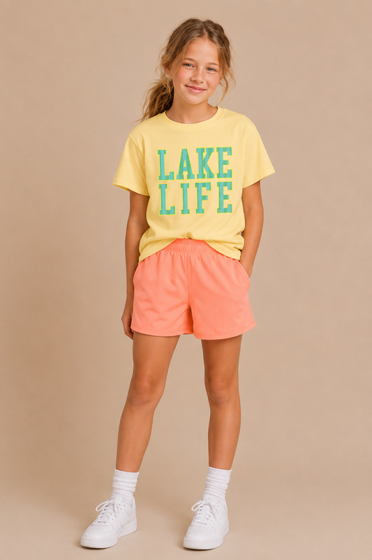 A young girl smiles in a Tiny Tot Bundles Lake Life Youth Tee, featuring green and blue lettering, paired with peach shorts, white crew socks, and white sneakers against a neutral beige background.