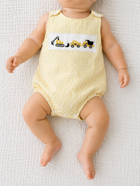 A baby in the Tiny Tot Bundles Sunshine Builder Romper, featuring yellow and white stripes with embroidered construction vehicles, lies on a textured white blanket.