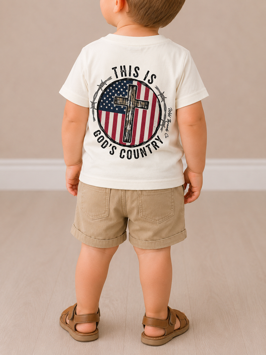 A young child wears the Wild Threads Co God's Country T-Shirt, featuring a cross and American flag graphic with the words THIS IS GOD'S COUNTRY, paired with khaki shorts and brown sandals on a light wood floor.