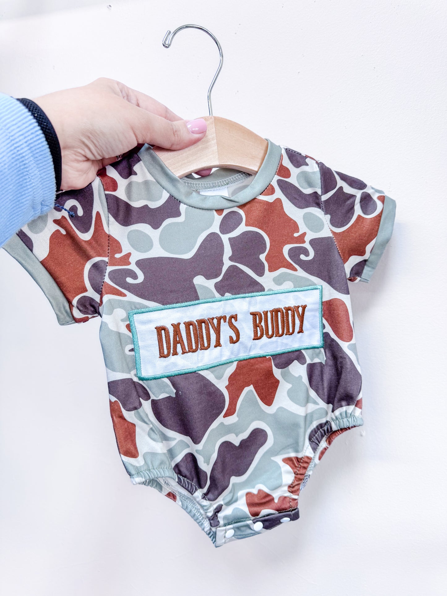 A hand holds up the Tiny Tot Bundles Daddy's Buddy Camo Romper—featuring a brown, green, and gray camo pattern and an orange and brown "DADDY'S BUDDY" patch—on a wooden hanger against a white wall.