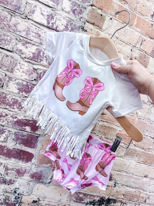 A hand displays the Tiny Tot Bundles Fringe Coquette Southern Gal Set—a white fringed top with pink cowboy boots and bows, paired with matching patterned shorts—against a weathered brick wall.