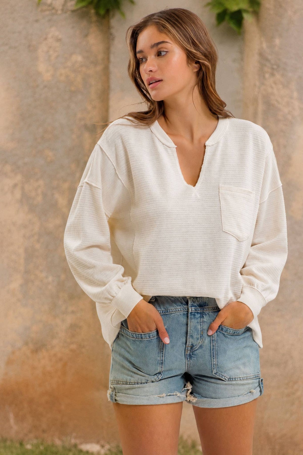 A woman stands outdoors against a textured wall, wearing the Blu Pepper Ivory Knit Long Sleeve Top paired with denim shorts, her hands in her pockets as she looks to the side.