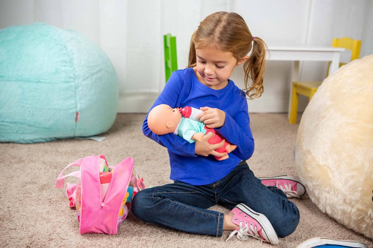 A young girl sits on a carpet, feeding her baby doll with a bottle from the Toysmith My Sweet Baby Deluxe Baby Ensemble 12-Piece Doll Playset. She wears a blue shirt and pink sneakers. Beside her is a pink bag filled with doll accessories, perfect for imaginative pretend play. The room features soft, colorful furniture in the background.