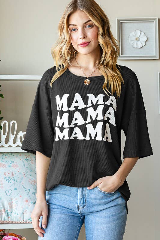 A woman with wavy blonde hair is wearing the HEIMISH Solid Ribbed MAMA Shirt, which features “MAMA” repeated in white letters on a black background. She stands indoors with one hand in her jeans pocket and smiles gently at the camera.