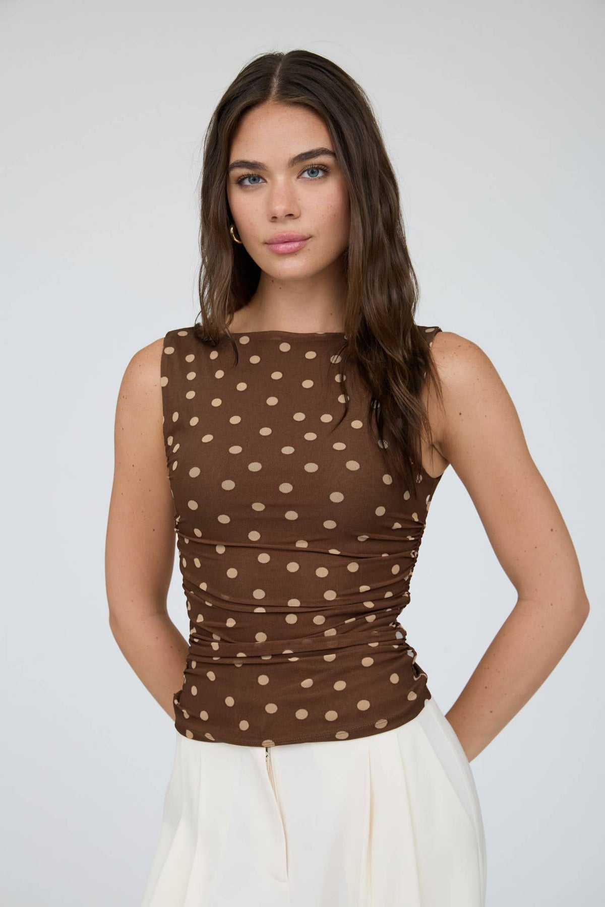 A woman with long brown hair wears The Workshop Studio’s Brown Polka Dot Boat Neck Top and a white skirt, standing against a plain light background.