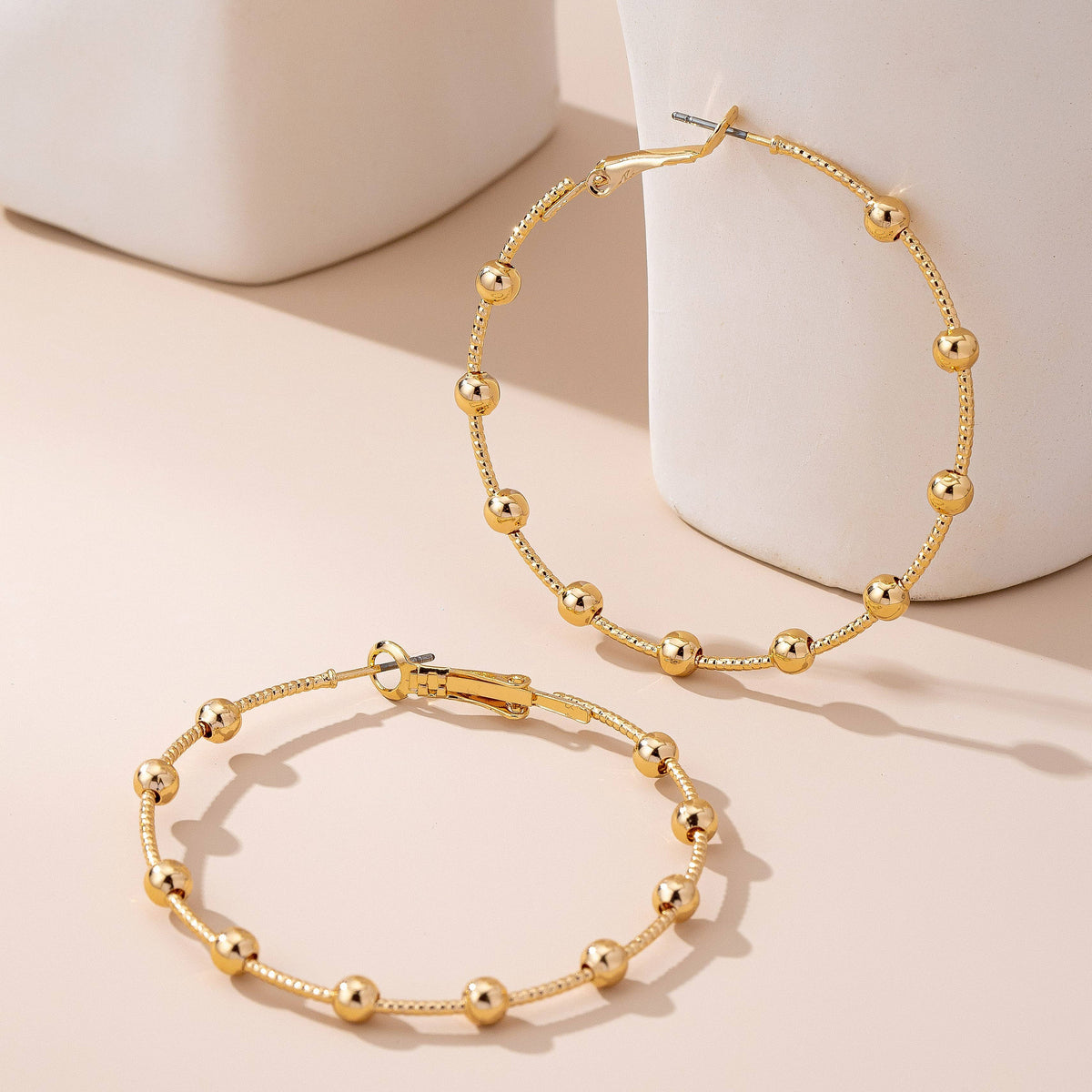 Two Gold Ball Beaded Hoop Earrings from JOSSLYN by wall to wall are arranged on a beige surface next to white ceramic objects, casting soft shadows.