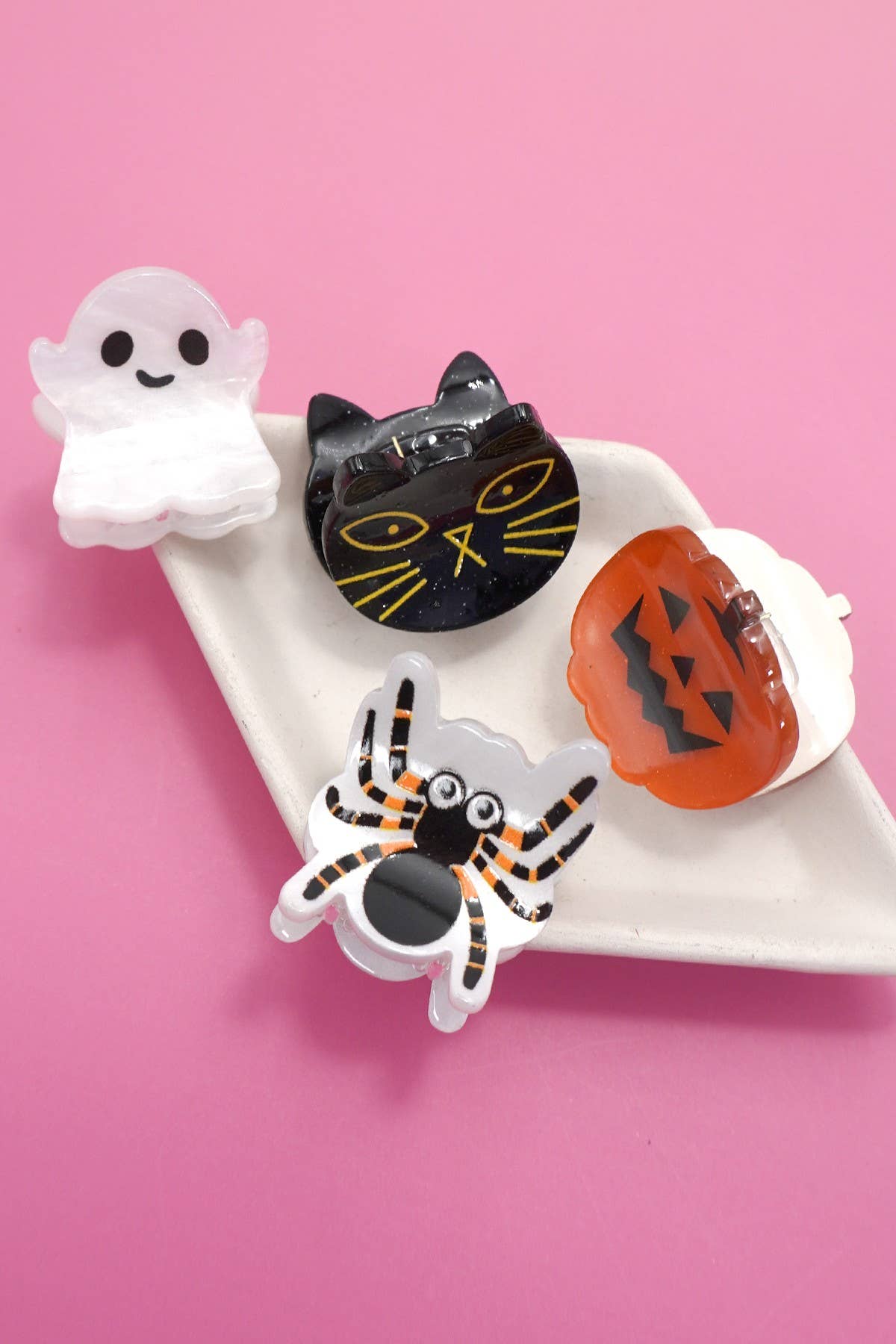 Four Halloween Ghost Theme Mini Hair Clips by JOSSLYN by wall to wall—a ghost, black cat, pumpkin, and spider—are displayed on a white dish against a pink background.