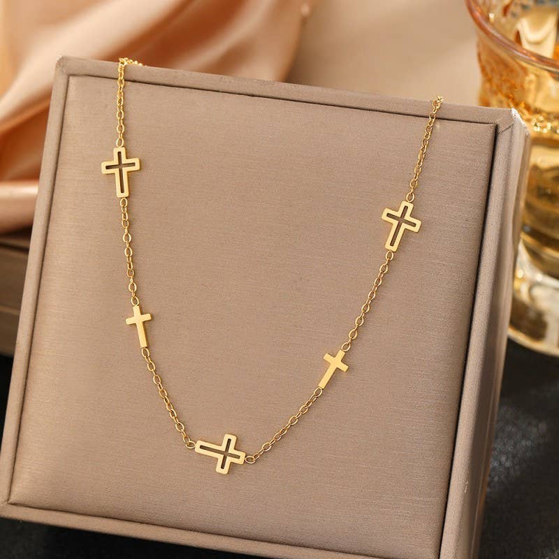 JOSSLYN by wall to wall’s Dainty Cross Necklace, featuring five delicate cross pendants, is displayed on a beige jewelry stand with a gold and glass ornament and a satin cloth in the background.