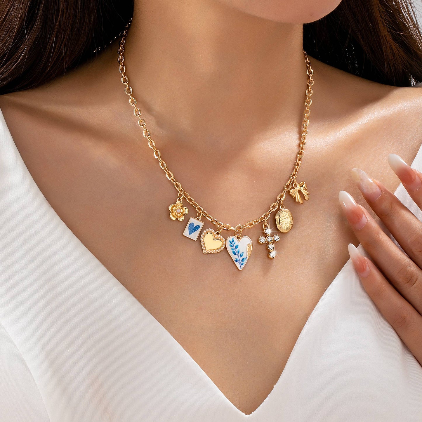 A woman wearing the JOSSLYN by wall to wall Blue Vintage Coquette Charm Necklace, featuring gold, heart, and floral charms with blue, white, and gold designs, over a white v-neck top.