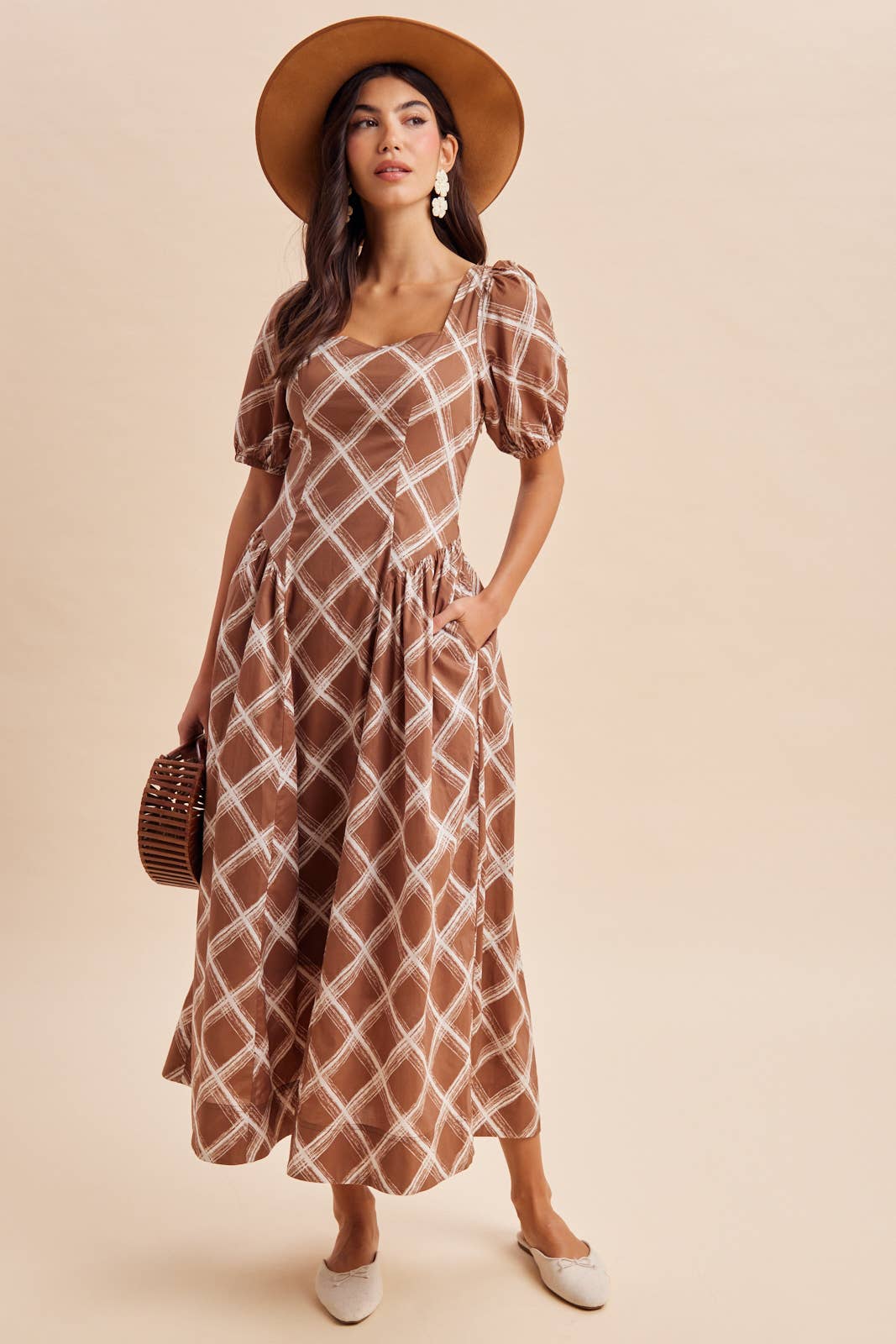 A person wearing an In Loom Plaid Square Neck Maxi Dress in brown and white plaid with puffed sleeves and side pockets, paired with a wide-brimmed hat, flat shoes, and a small wooden handbag.