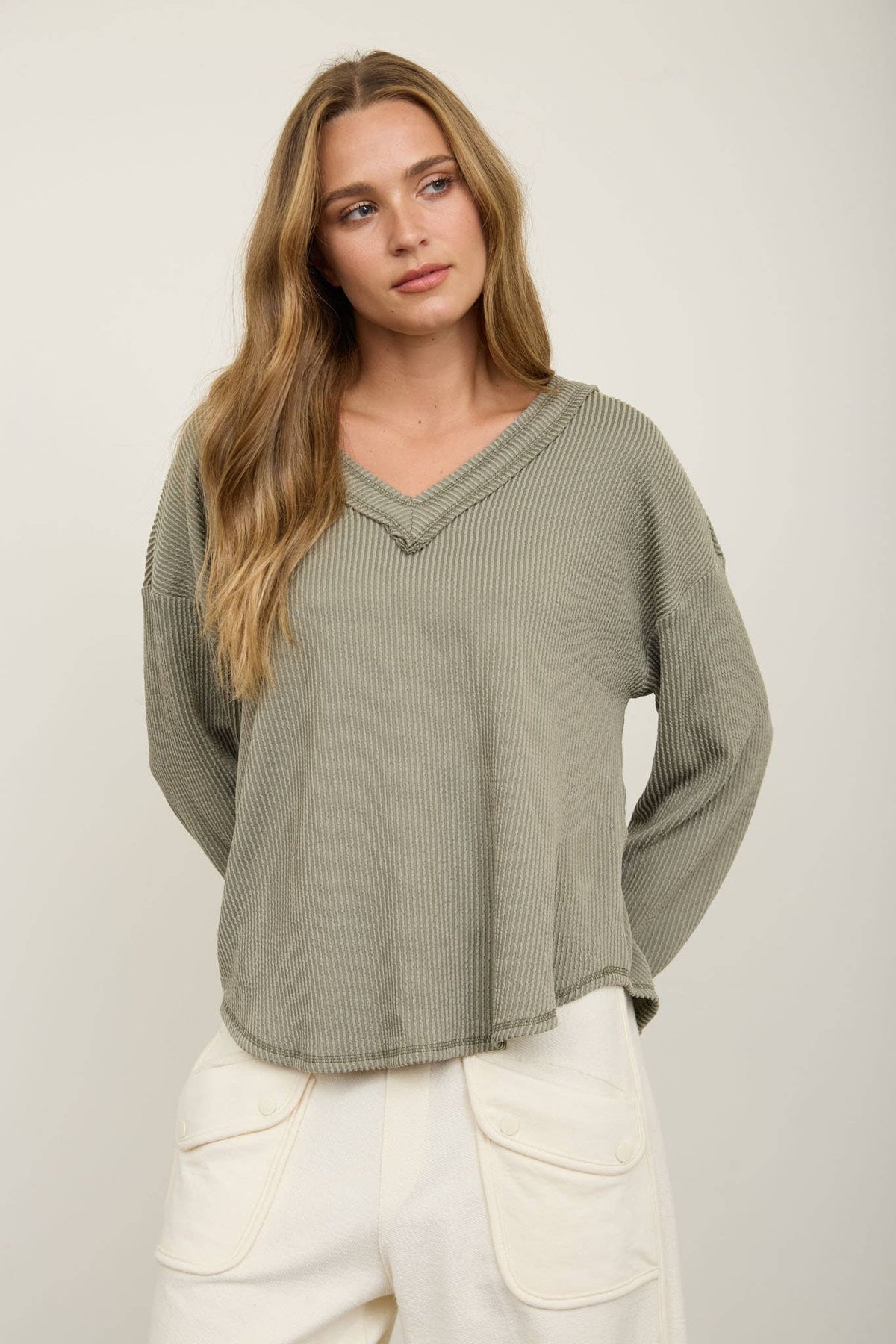 A woman with long wavy hair is wearing the Blu Pepper Olive V-Neck Long Sleeve Top—an easy, loose-fitting, ribbed sweater—paired with white pants featuring large pockets, standing against a plain light background.
