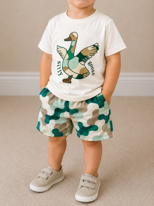 A young child wears the Tiny Tot Bundles Waddlin’ West Short Set—featuring a white t-shirt with a cartoon goose labeled "silly goose" and green camo shorts—paired with beige sneakers, standing on a light carpet.