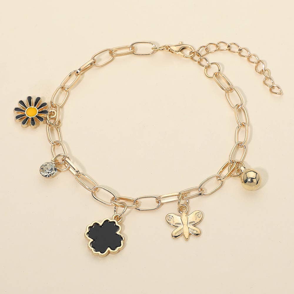The Charm Station Paperclip Chain Bracelet by Sensibling Corp. features five dangling charms—a daisy, clear gemstone, black flower, butterfly, and gold sphere—displayed on a light beige background.