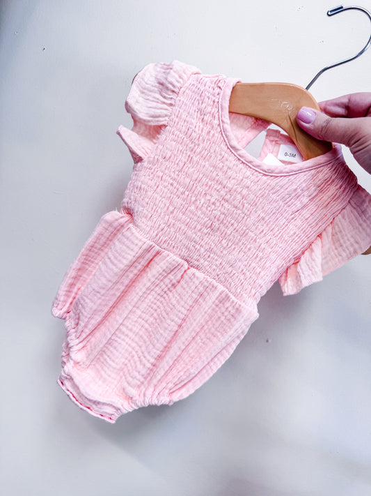 A hand holds the Tiny Tot Bundles Peach Petal Romper—a light pink, short-sleeved baby outfit with a smocked bodice and ruffled sleeves—against a plain white background.