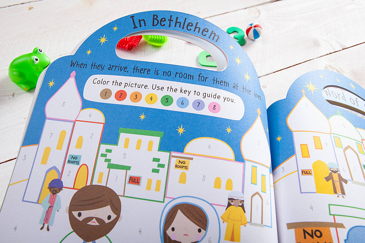 Here’s a revised sentence using the provided product data: The Story of Christmas Activity Book by BroadStreet Publishing Group, LLC features an “In Bethlehem” page with vibrant illustrations of people and buildings, a color-by-number key, and toys in the background on a white wooden surface.