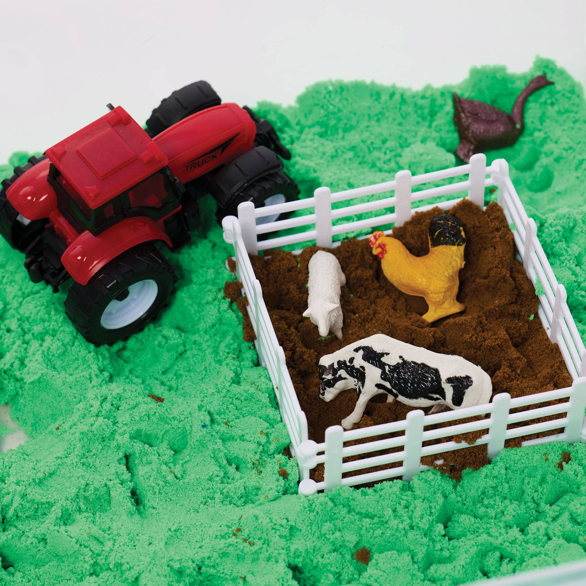 The Sensory Bin Farm Activity Kit for Kids from Faber-Castell / Creativity for Kids features a toy red tractor resting on green kinetic sand inside a sensory bin, alongside a fenced pen with toy farm animals—including a cow, pig, and chicken—on brown sand. A toy horse appears in the background, encouraging children to explore and develop fine-motor skills through imaginative play.