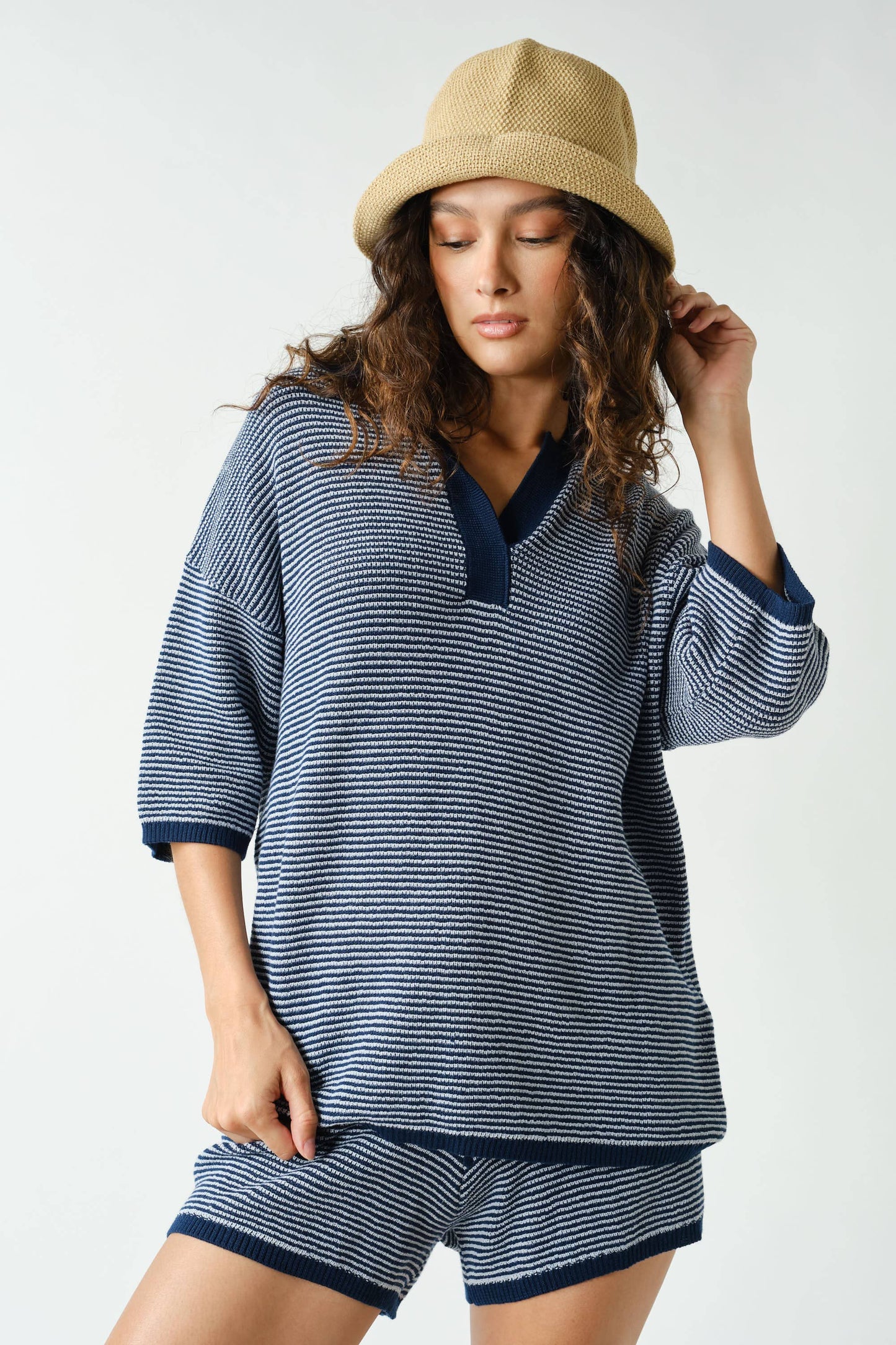 A woman wearing the Mono B Navy Coastal Breeze Knit Set—featuring a blue and white striped collared top and matching shorts—stands against a plain background, looking down with one hand touching her beige hat.