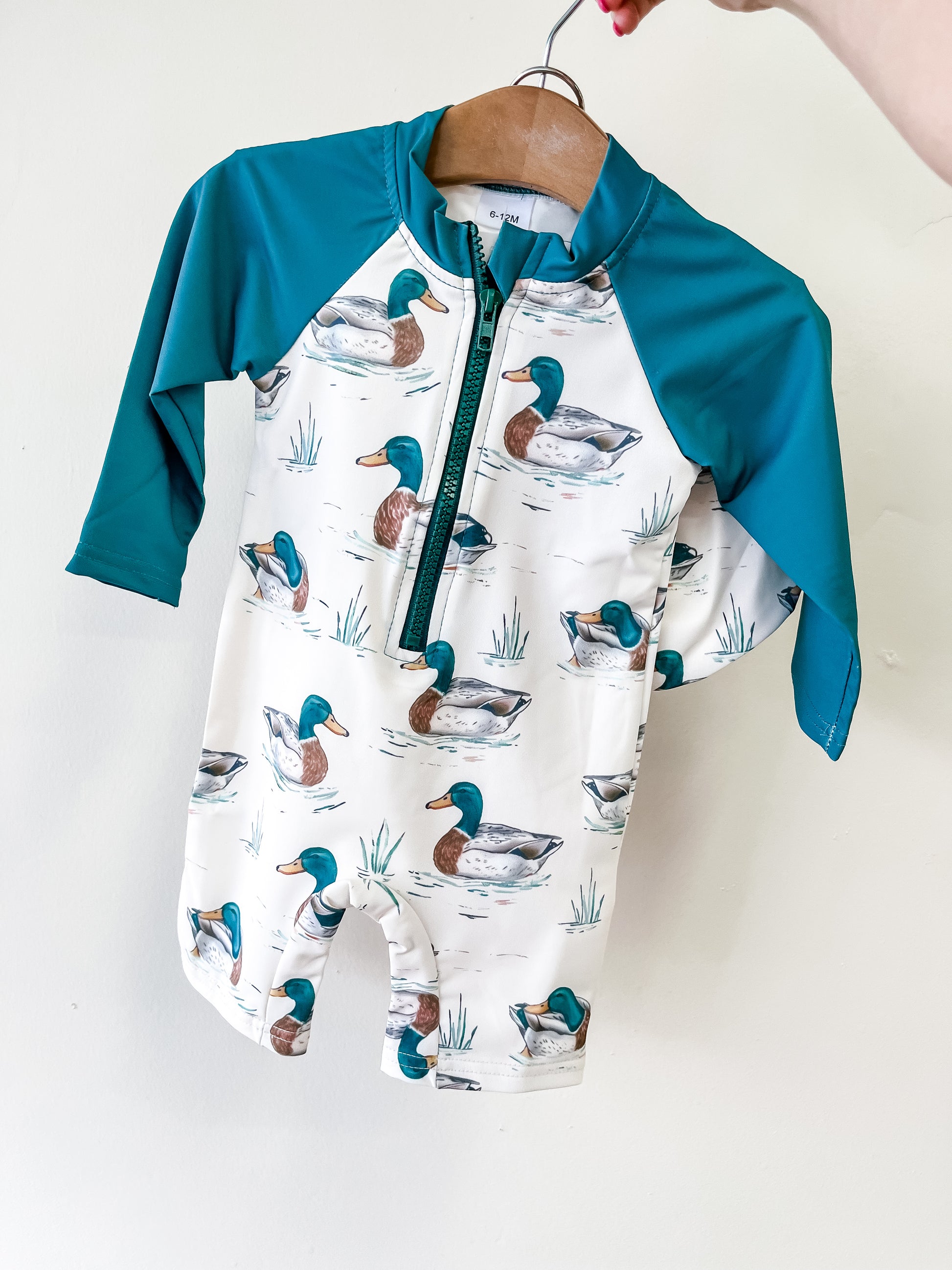 The Little Duckling Swim Romper by Tiny Tot Bundles is a long-sleeve baby swimsuit with teal sleeves and a white body decorated with swimming duck patterns, shown on a wooden hanger against a white background.