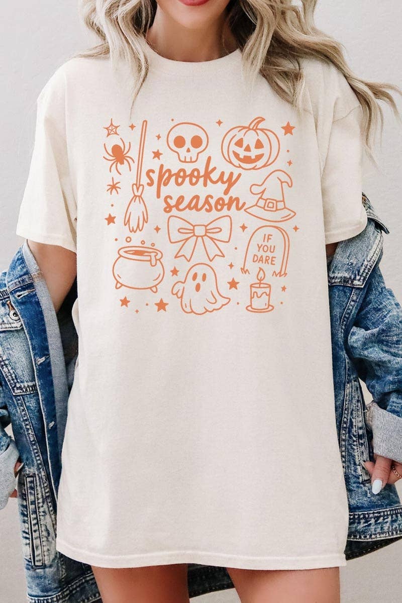 A woman wears the COLORBEAR Halloween Doodle Graphic Heavy Cotton Tee, a cream-colored shirt featuring orange Halloween-themed doodles—ghost, pumpkin, broom, skull, witch hat—and the phrases “spooky season” and “if you dare.” The tee is styled with a denim jacket draped over her shoulders.