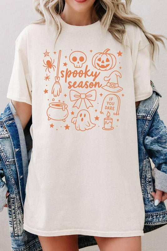A woman wears the COLORBEAR Halloween Doodle Graphic Heavy Cotton Tee, a cream-colored shirt featuring orange Halloween-themed doodles—ghost, pumpkin, broom, skull, witch hat—and the phrases “spooky season” and “if you dare.” The tee is styled with a denim jacket draped over her shoulders.