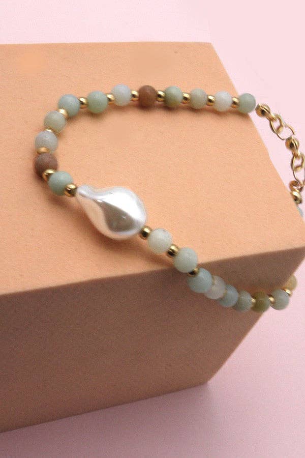 The Stone Beaded Pearl Bracelet by JOSSLYN by wall to wall features light green and gold beads with a large teardrop-shaped white pearl centerpiece, showcased on a beige box against a pink background.