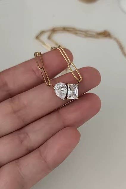 A hand holds the Gold Rhin Necklace by JOSSLYN by wall to wall, featuring two large clear gemstone pendants—one pear-shaped and one square—on a gold chain. The background is blurred.
