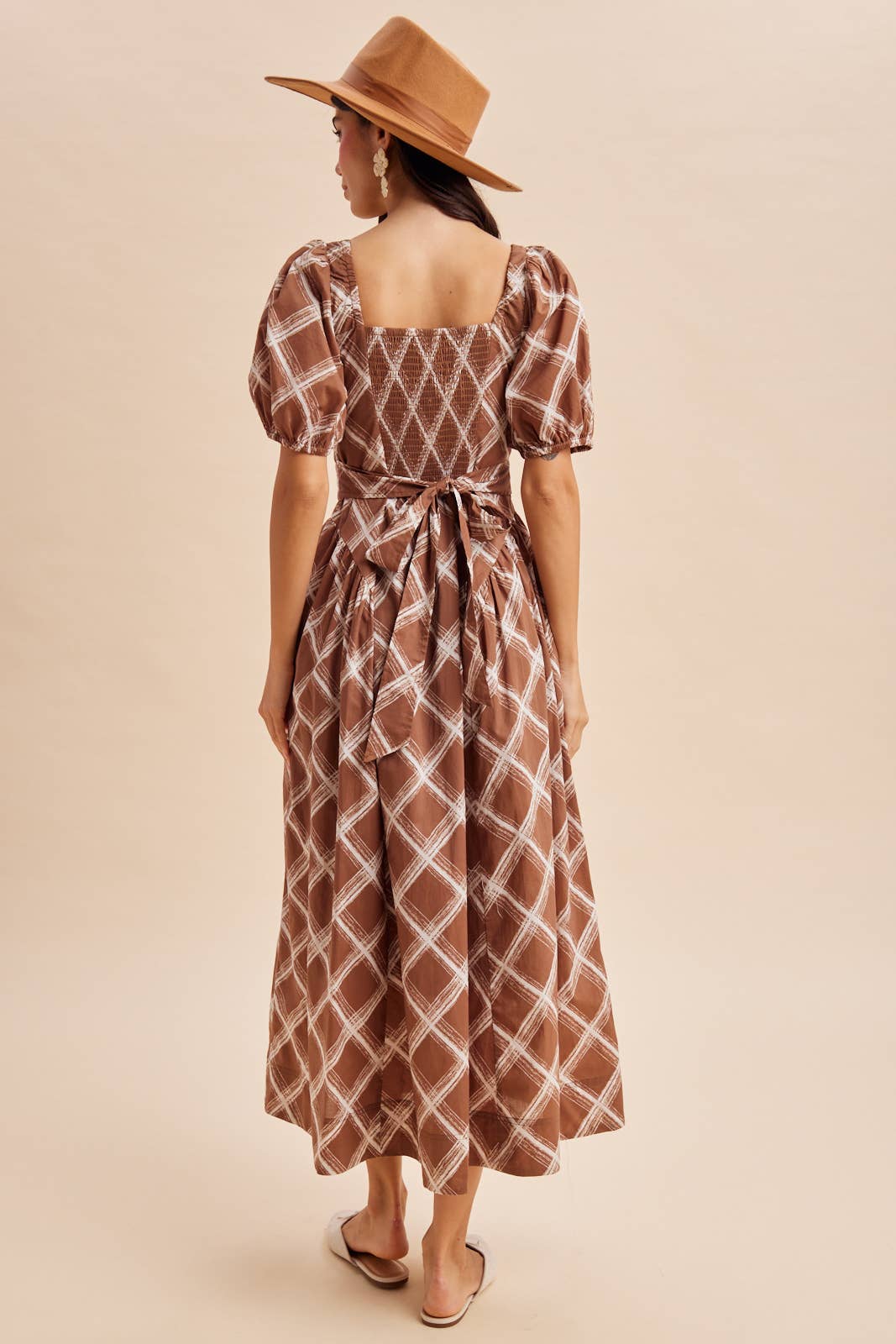 Back view of a person wearing an In Loom Plaid Square Neck Maxi Dress in brown and white, featuring a square neckline, puffed sleeves, and a tied waist, paired with a brown hat.