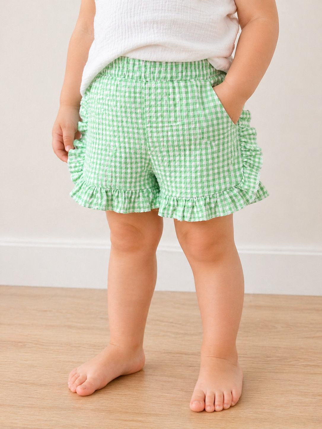 A young child stands barefoot on a wooden floor, wearing Tiny Tot Bundles Picnic Day Gingham Shorts in green and white, paired with a white top, hands in the shorts pockets. The child's face is not visible.