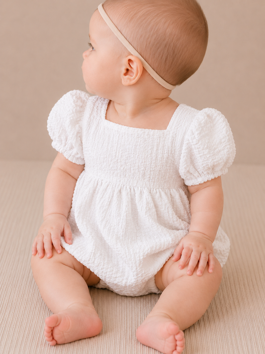 A baby in the Tiny Tot Bundles Sweet Peony Romper sits on a neutral surface, looking to the side. The baby wears a beige headband, with hands on their legs and bare feet.