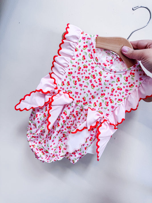 A hand displays the Tiny Tot Bundles Berry Bow Belle Romper, featuring a cherry print, pink ruffled trim, red zigzag edging, and two large bows on the front against a white background.