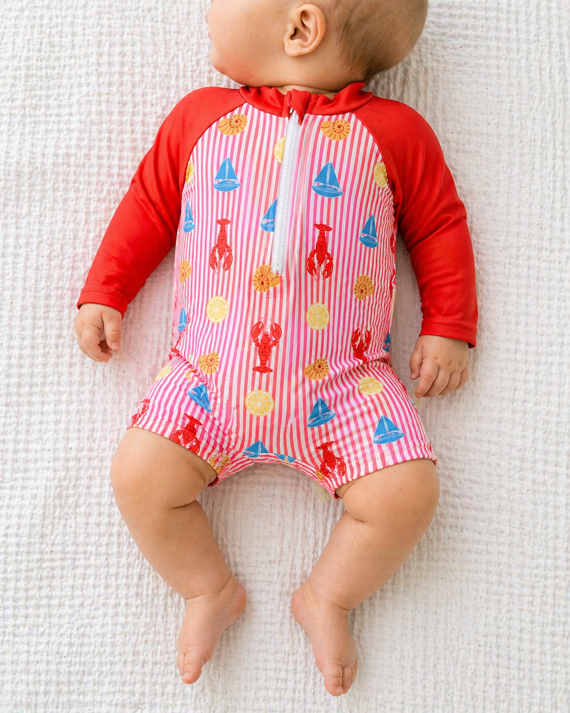 A baby in the Lobster Lagoon Swimsuit by Tiny Tot Bundles, featuring red sleeves and playful lobster, sun, and sailboat prints, lies on a white textured blanket.