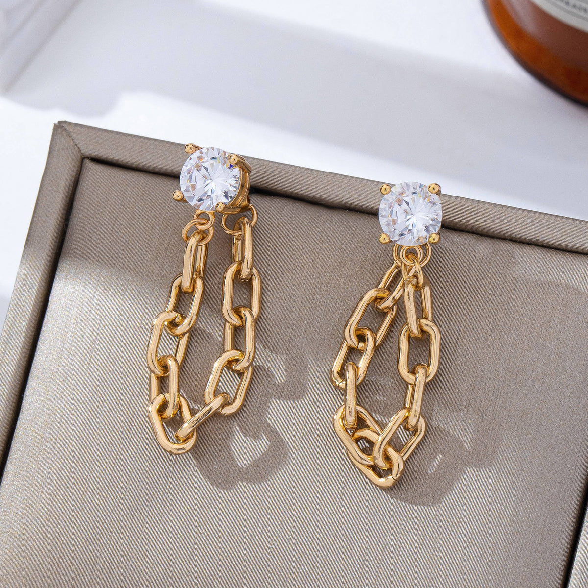 JOSSLYN by wall to wall Stud Hoop and Drop Link Earrings featuring clear round gemstone studs and gold chain links, displayed on a beige jewelry box in direct natural light.