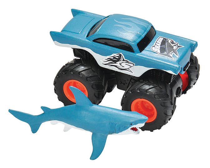 The Wild Republic Adventure-Mini Truck Shark is a blue toy monster truck with large black wheels, orange hubs, and shark-themed graphics, shown next to a small blue plastic shark figure with an open mouth.