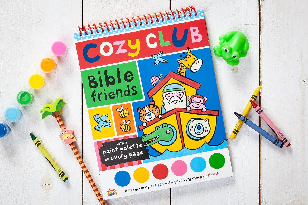 A vibrant children’s art pad, the Cozy Club Bible Friends Art Pad Book by BroadStreet Publishing Group, LLC, featuring animal illustrations, paint, crayons, a toy frog, and a paint palette with brush on a white wooden surface.
