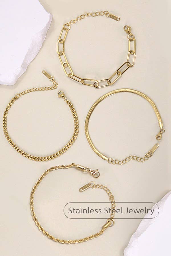 Four Dainty Gold Bracelets by JOSSLYN by wall to wall, each featuring a unique chain style, are arranged on a beige surface and surrounded by pieces of white stone. A label reads Stainless Steel Jewelry.